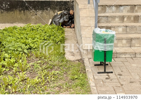 A green garbage container is installed on the sidewalk next to the stairs, next to it lies a black garbage bag covered with building bricks in the corner of the house A green garbage container is installed on the sidewalk next to the stairs, next to it lies a black garbage bag covered with building bricks in the corner of the house 114933970