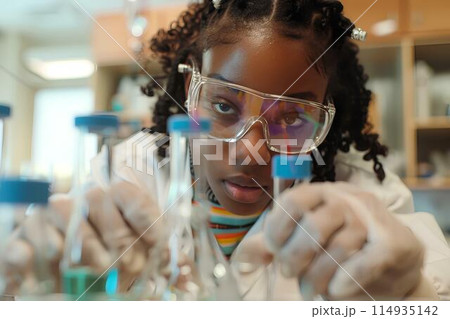 Black girl doing a science experiment in school, at a desk with test tubes and beakers, wearing glasses, with a chalkboard background showing a globe and educational posters 114935142