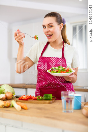 Portrait of woman standing in home kitchen and tasting vegetable salad 114935145