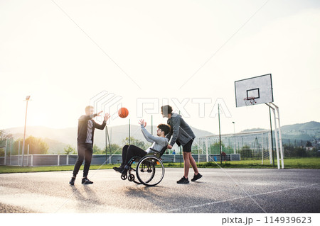 Disabled young man in a wheelchair playing basketball with his friends. Teamwrok and male friendship. Disabled young man in a wheelchair playing basketball with his friends. Teamwrok and male friendship. 114939623