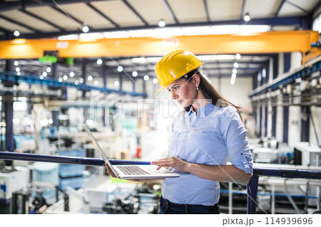 Female project manager standing in modern industrial factory. Manufacturing facility with robotics, robotic arms and automation. Storing products and materials in warehouse. 114939696
