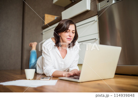 Mature woman working from homeoffice. Woman paying bills online, lying on floor. 114939848