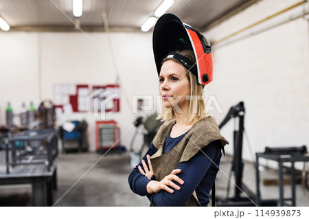 Beautiful blonde woman works as a welder in workshop, operating welding machine, wearing protective clothing and a welding mask. Beautiful blonde woman works as a welder in workshop, operating welding machine, wearing protective clothing and a welding mask. 114939873