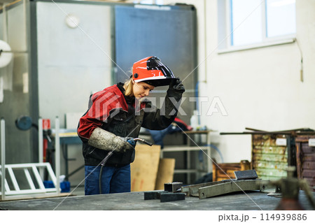 Beautiful blonde woman works as a welder in workshop, operating welding machine, wearing protective clothing and a welding mask. 114939886