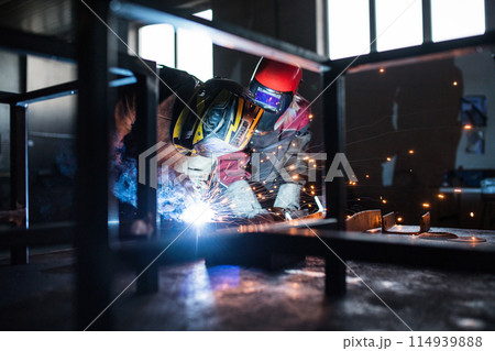 Blonde woman works as a welder in workshop, operating welding machine, wearing protective clothing and a welding mask. Two welders helpign each other. 114939888