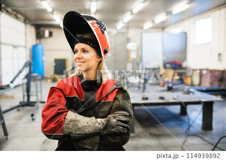 Beautiful blonde woman works as a welder in workshop, operating welding machine, wearing protective clothing and a welding mask. Beautiful blonde woman works as a welder in workshop, operating welding machine, wearing protective clothing and a welding mask. 114939892