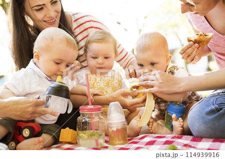 Mothers and babies enjoying group picnic outdoor in park, sitting on picnic blanket and preparing food and drinks. 114939916