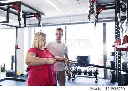 Overweight woman exercising on gym mat in gym. Personal trainer couching her and helping her. 114939920