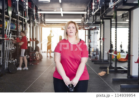 Overweight woman exercising in gym, using kettlebell. Personal trainer couching her and helping her. 114939954