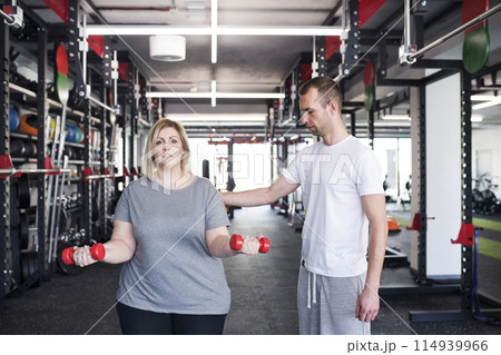Overweight woman exercising in gym, using dumbbells. Personal trainer couching her and helping her. Overweight woman exercising in gym, using dumbbells. Personal trainer couching her and helping her. 114939966