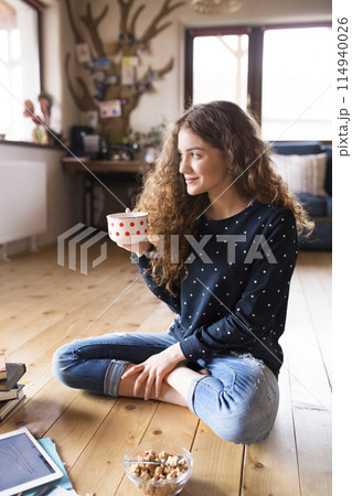 Female student sitting on floor, reading books, studying for final exam at university. Beautiful student at home, drinking coffee. 114940026