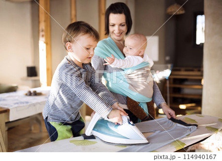 Mother carrying small baby in wrap, older son helping with ironing. Family doing household chores. 114940167