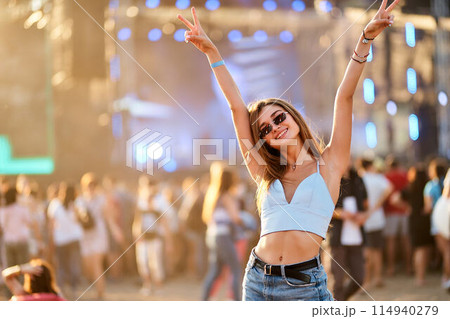 Happy young woman at outdoor music festival raises peace signs, enjoys summer event on sunny day. Crowd dances, socializes at beach concert. Fashionable festivalgoer lives up moment, carefree vibes. 114940279