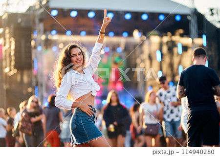 Happy young woman enjoys summer music festival, dancing with hand up, stage with lights in background. Casual fashion, denim shorts, carefree vibe at outdoor concert with crowd. Sunset atmosphere. Happy young woman enjoys summer music festival, dancing with hand up, stage with lights in background. Casual fashion, denim shorts, carefree vibe at outdoor concert with crowd. Sunset atmosphere. 114940280