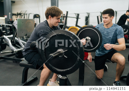 Motivated beginner sportsman hard time working out in gym with personal trainer at gym. Trainer helping beginner sportsman for correct doing Scott Curls with barbell, lifting up EZ-bar. Motivated beginner sportsman hard time working out in gym with personal trainer at gym. Trainer helping beginner sportsman for correct doing Scott Curls with barbell, lifting up EZ-bar. 114941151