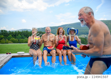 Group of cheerful seniors sitting on edge of pool with legs in water, eating watermelon. Elderly friends spending hot day by swimming pool, sunbathing. Group of cheerful seniors sitting on edge of pool with legs in water, eating watermelon. Elderly friends spending hot day by swimming pool, sunbathing. 114941507