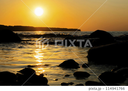 Sun setting over ocean, casting warm glow on rocks in foreground 114941766