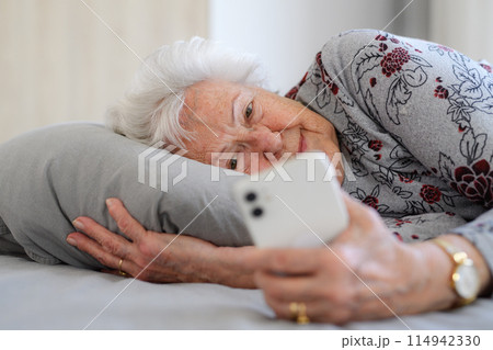 Senior woman lying in bed, looking at smartphone and smiling. Older woman using technology, digital skill and literacy. 114942330