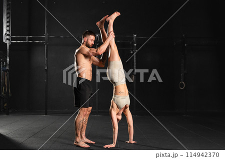 Muscular woman doinghandstand exercise in gym, man helping her. Routine workout for physical and mental health. 114942370