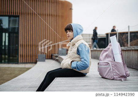 Sad girl alone, sitting in front of school building, waiting for parents. Young schoolgirl with no friends, feeling lonely. Sad girl alone, sitting in front of school building, waiting for parents. Young schoolgirl with no friends, feeling lonely. 114942390