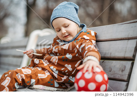 Cute toddler girl eating lunch in park, sitting on bench. Picnic in park during cold spring day. 114942396