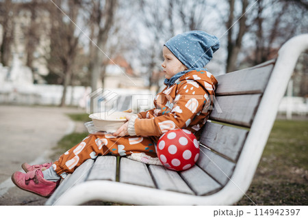 Cute toddler girl eating lunch in park, sitting on bench. Picnic in park during cold spring day. 114942397
