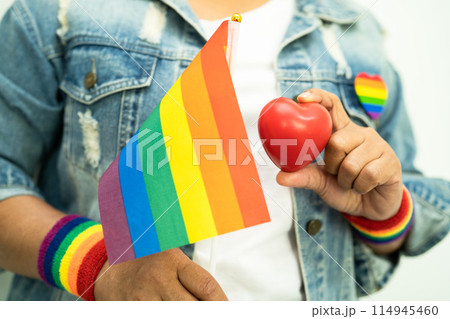 Asian lady wearing rainbow flag wristbands and hold red heart, symbol of LGBT pride month celebrate annual in June social of gay, lesbian, bisexual, transgender, human rights. Asian lady wearing rainbow flag wristbands and hold red heart, symbol of LGBT pride month celebrate annual in June social of gay, lesbian, bisexual, transgender, human rights. 114945460