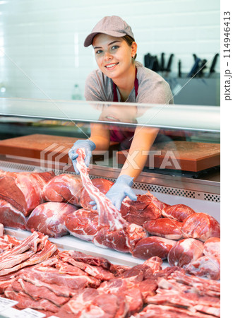 Positive young saleswoman vendor in apron and disposable gloves shows meat tenderloin Positive young saleswoman vendor in apron and disposable gloves shows meat tenderloin 114946413