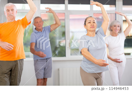 Happy aged woman in sportswear confidently leading ballet class with dancers following movements in dance studio Happy aged woman in sportswear confidently leading ballet class with dancers following movements in dance studio 114946453