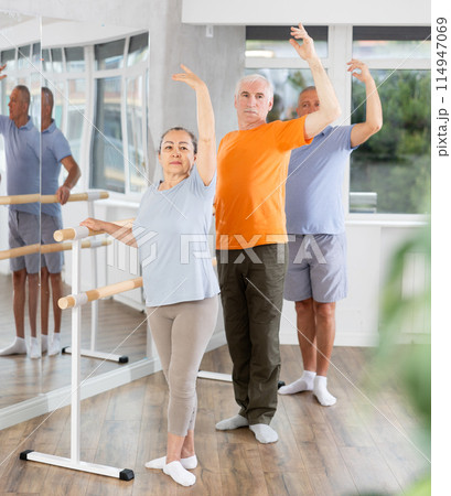 Ordinary elderly people stand in third position near ballet barre during group training in dance studio Ordinary elderly people stand in third position near ballet barre during group training in dance studio 114947069