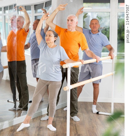 Cheerful active elderly woman training various ballet poses during group classes with ballet dancers at barre in choreography studio 114947087