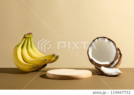 Advertising photo with a wooden pedestal in round shaped displayed on brown table next to some coconut and a bunch of banana, over light yellow background. Vacant space for design or adding text 114947752