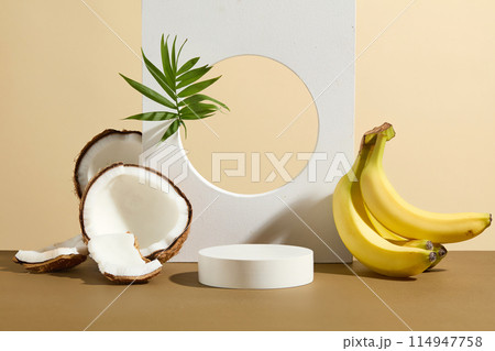 The white backdrop has a hole in center striking with a green tree branch, white geometric podium next to a bunch of yellow banana and some pieces of coconut featured on light yellow background 114947758