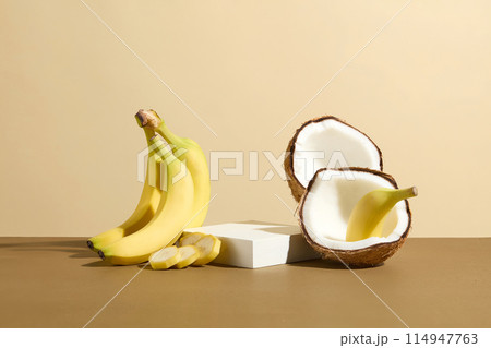 White showcase platform in rectangular shape beside banana and coconut composition featured on brown countertop over light yellow background. Coconuts and bananas are both high in potassium 114947763