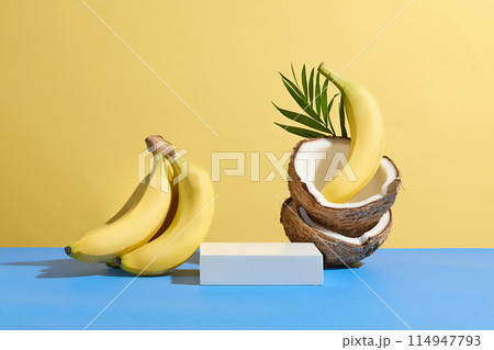 Tropical fruit concept photo with yellow background and blue countertop, an empty pedestal in rectangular shaped placed between a bunch of banana and a pile of dry coconut Tropical fruit concept photo with yellow background and blue countertop, an empty pedestal in rectangular shaped placed between a bunch of banana and a pile of dry coconut 114947793
