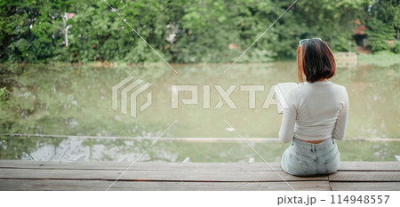 A woman is sitting on a wooden bench by a lake, reading a book. The scene is peaceful and serene, with the woman enjoying the quiet and calm atmosphere 114948557