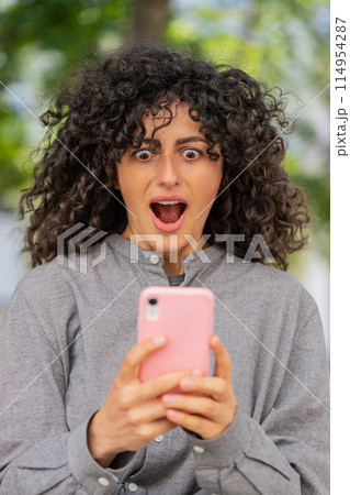 Excited happy Caucasian young woman use smartphone celebrating win good message news in city street Excited happy Caucasian young woman use smartphone celebrating win good message news in city street 114954287