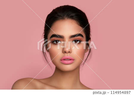 Young woman with dark hair, pink lips, and a poised expression, set against a soft pink background 114954897