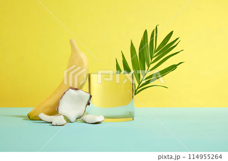 A empty glass podium placed in center with coconut pieces, banana and green coconut leaf decoration on blue table top over yellow background. Frontal shot photography with vacant space for design 114955264