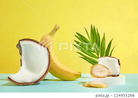 A yellow banana and a coconut piece leaning on each other, next to a round podium contains banana slices and coconut above, decorated with green coconut leaf behind. Copy space, front view 114955266