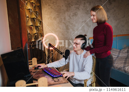 Young attractive wife help husband to work. Handsome man sits at the table and work on computer. Man using professional microphone and headphones 114961611
