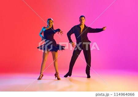 Dynamic shot of young man and woman dancing Zumba in motion in neon lighting against vibrant gradient background. Dynamic shot of young man and woman dancing Zumba in motion in neon lighting against vibrant gradient background. 114964066
