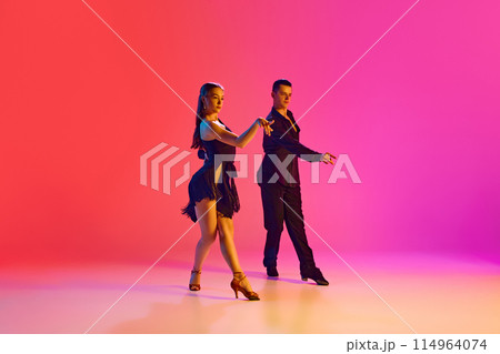 Graceful ballroom dancers in black stage costumes performing Cha Cha in neon lighting against vibrant gradient background. Graceful ballroom dancers in black stage costumes performing Cha Cha in neon lighting against vibrant gradient background. 114964074