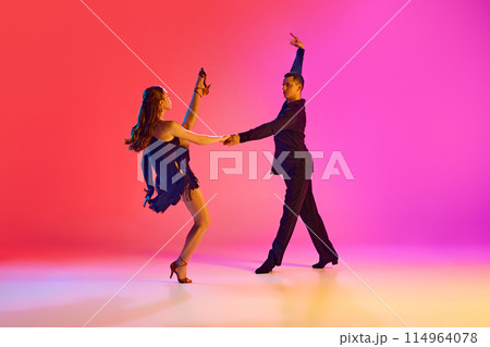 Professional ballroom dancers in black stages costumes performing in neon lighting against vibrant gradient background. 114964078