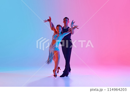Pair of ballroom dancers performing dramatic pose, with woman in shimmering dress and man in sleek black outfit in neon lighting against gradient background. 114964203
