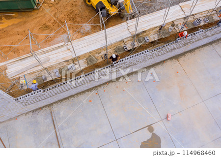 On construction site, mason makes concrete wall using cement blocks while standing on scaffolding On construction site, mason makes concrete wall using cement blocks while standing on scaffolding 114968460