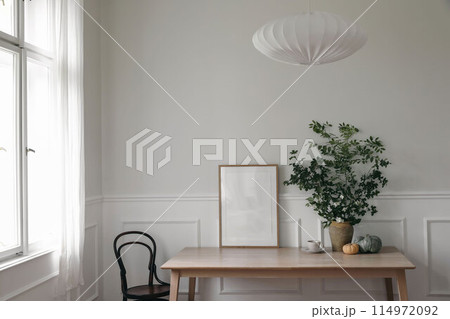 Modern Scandinavian living room interior. Wooden poster, picture frame mockup on office table, Green tree branches in vase, pumpkins, cup of coffee. Vintage chair near window. Stylish home decor. 114972092