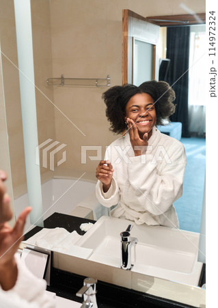 Vertical portrait of young African American woman looking in mirror enjoying skincare in morning, copy space 114972324