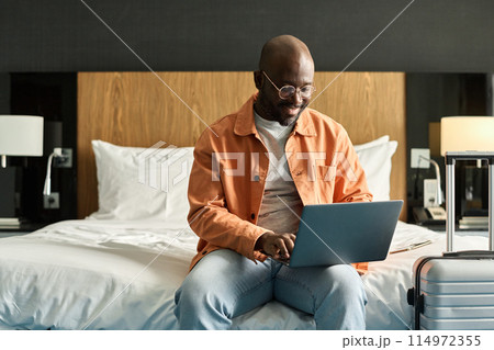 Portrait of smiling Black man using laptop sitting on bed in hotel room and researching accommodation facilities copy space 114972355
