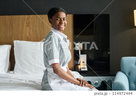 Side view portrait of young Black woman as housekeeper wearing uniform and smiling at camera sitting on bed in hotel room copy space 114972494